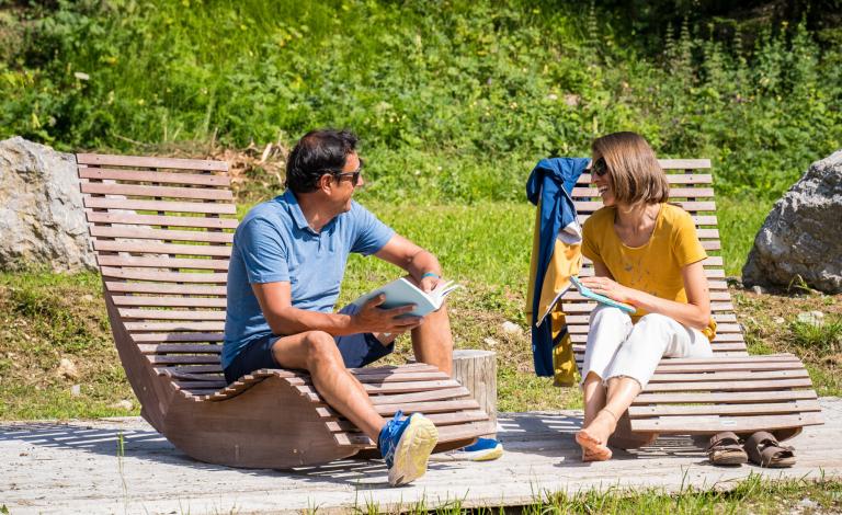 Due persone leggono all'aperto su sedie a sdraio di legno.