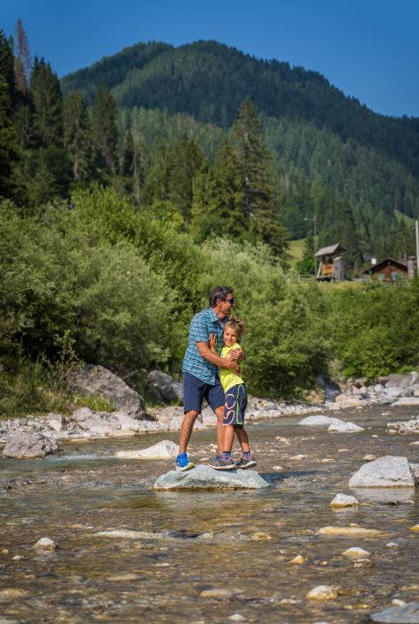 Padre e figlio abbracciati su una roccia nel fiume, circondati da natura rigogliosa.